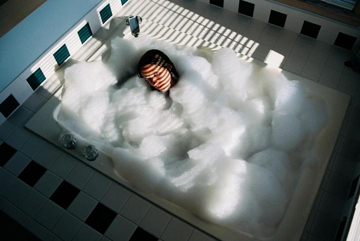 A woman is taking a bubble bath in a tiled bathroom with strong shadows from a window blind.