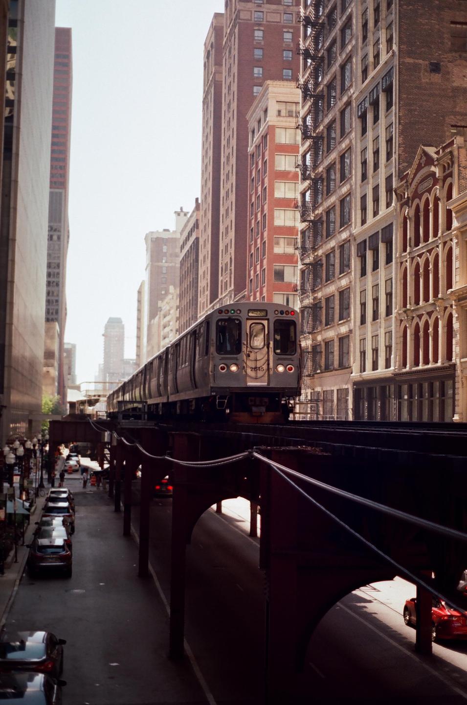 A train travels on an elevated track above a street lined with cars and tall buildings.