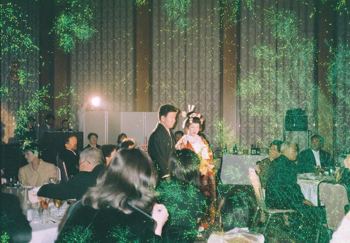 A couple in traditional Japanese wedding attire stands in a banquet hall with guests seated at tables, all covered in green light artifacts.