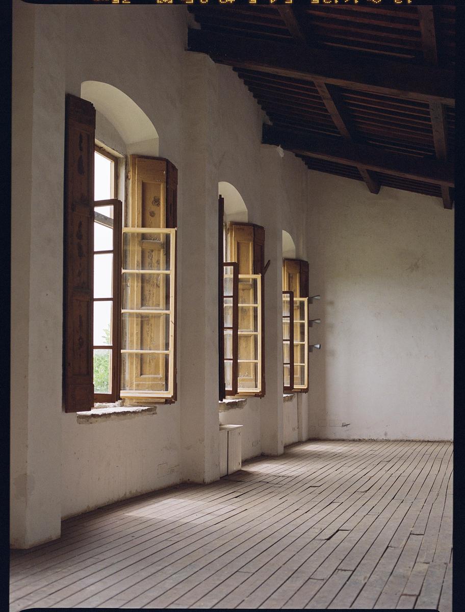 Sunlight streams through a row of open windows with wooden shutters onto a wooden floor.