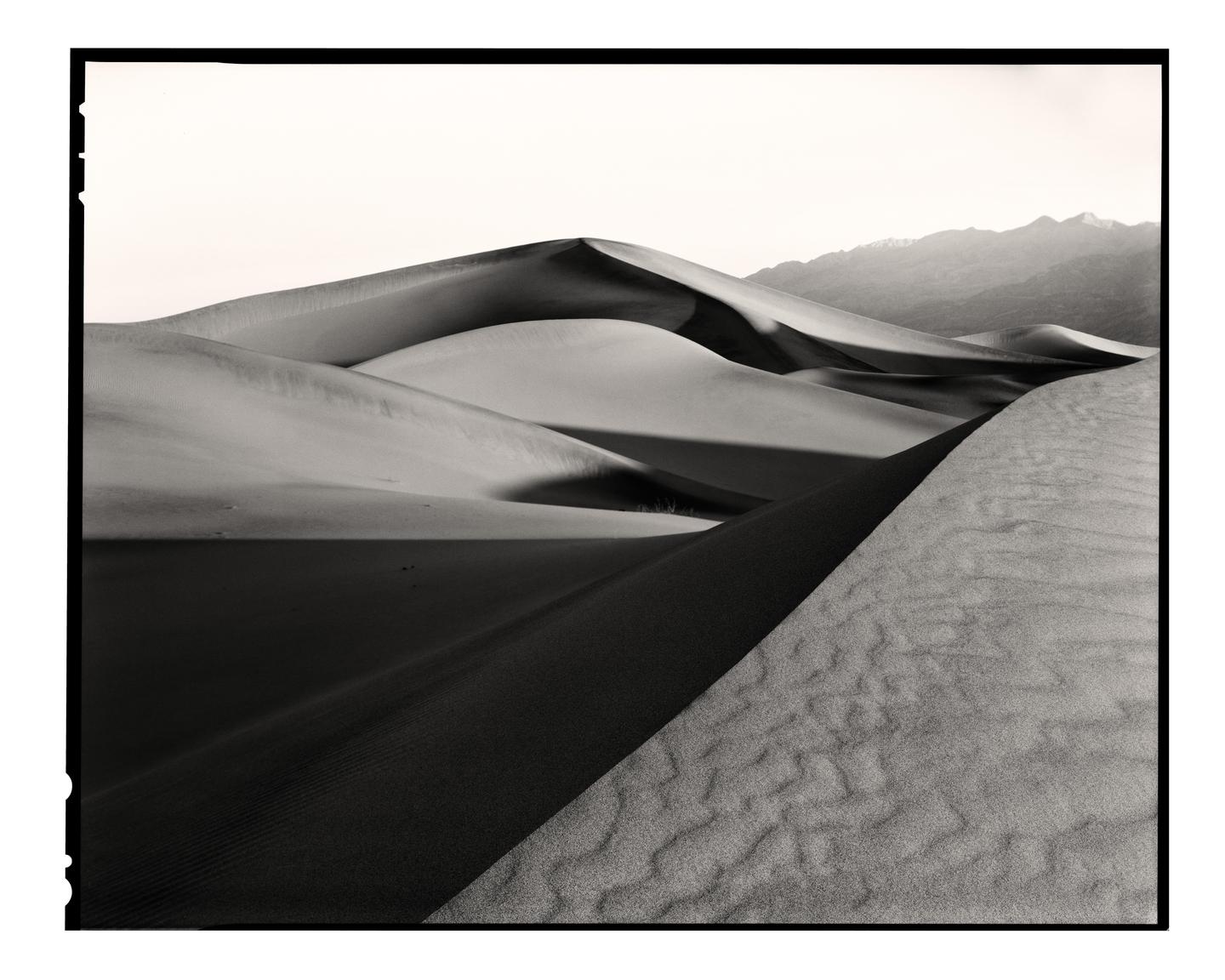 A black and white landscape shows sand dunes with shadows and mountains in the distance.