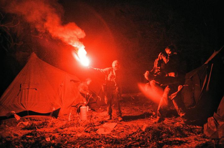 Three men are gathered around a red flare at night, with two tents visible in the background.