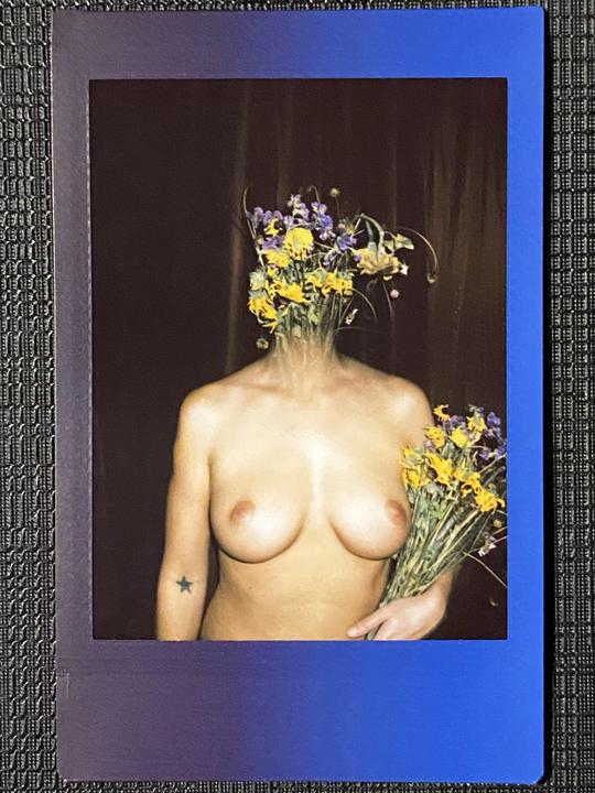 A nude woman holds a bouquet of flowers and has flowers covering her head.