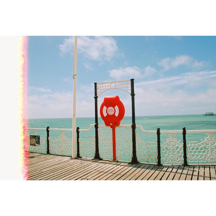 A red lifebuoy is mounted on a pier railing overlooking the ocean under a blue sky with clouds.