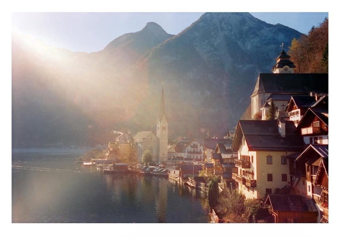A village with a prominent church steeple is nestled beside a lake, surrounded by mountains under a hazy sky with a strong sun flare.