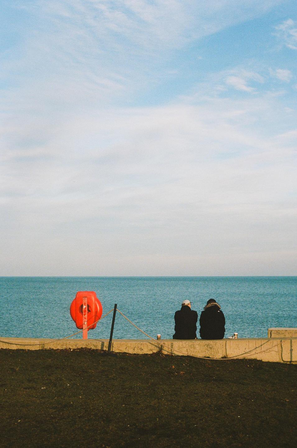 Two people sit on a concrete wall overlooking the ocean, with a red life preserver to their left.