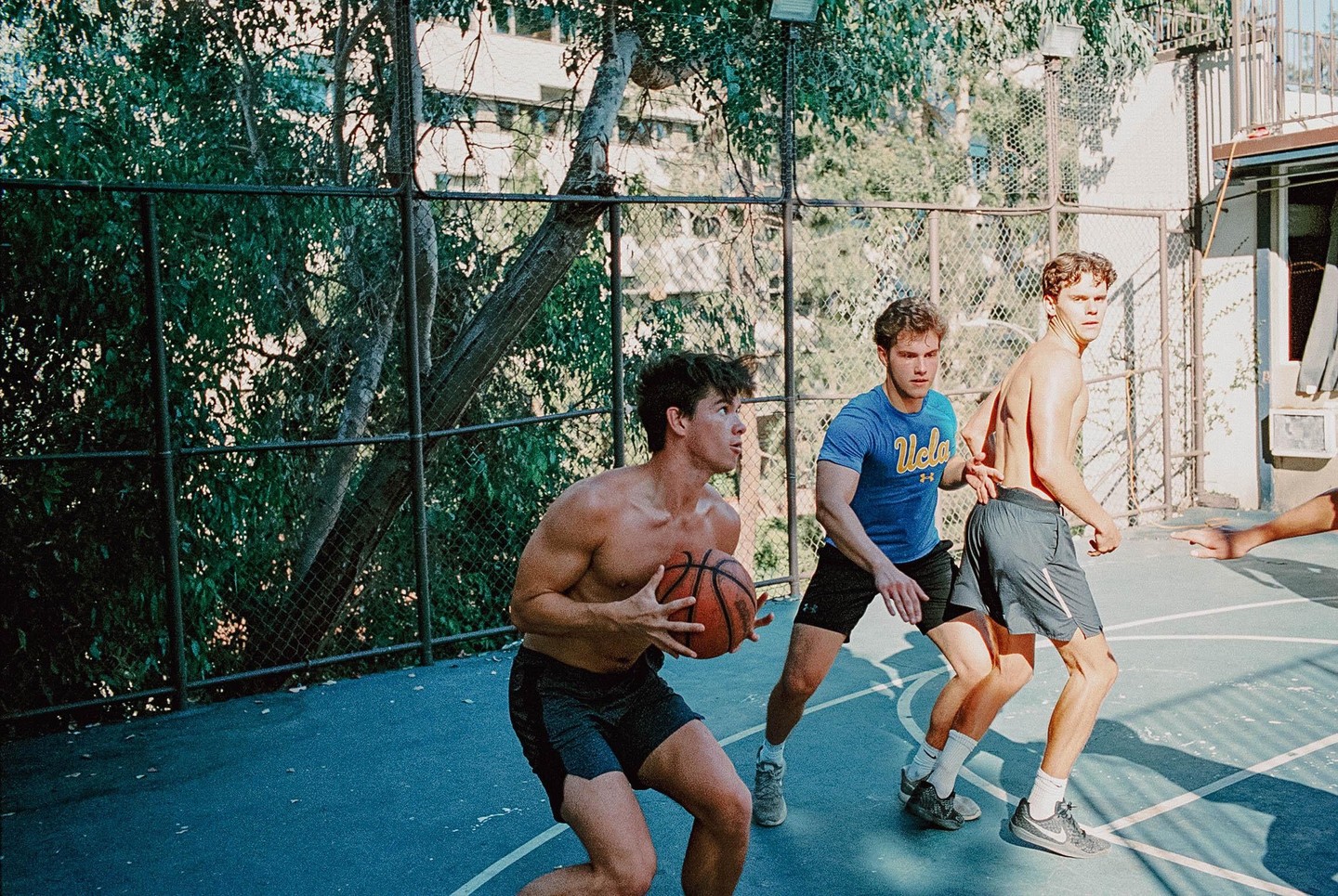 Three men play basketball on an outdoor court surrounded by a fence and trees.