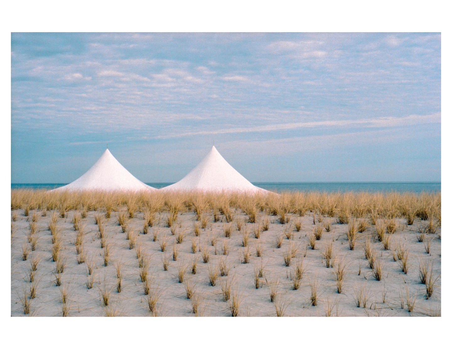 Two white conical tents are visible behind a sandy beach with sparse vegetation, with the ocean and a cloudy sky in the background.