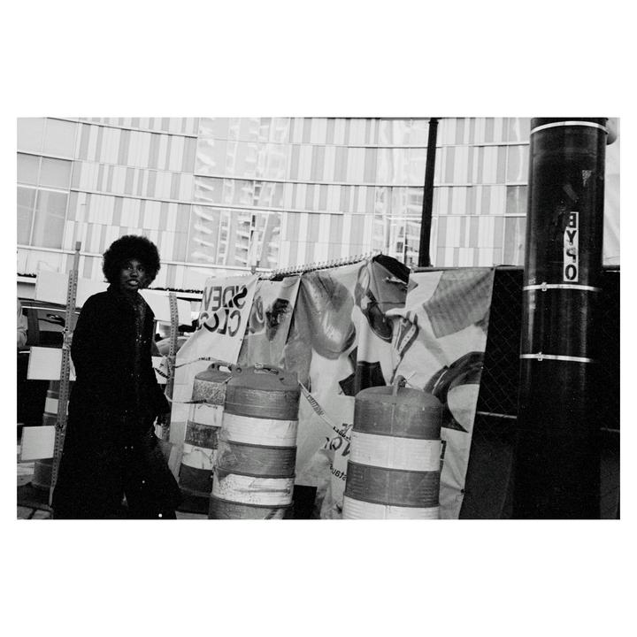 A man with an afro stands on a street next to construction barriers and a building.