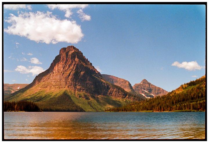 A large mountain rises from the edge of a lake under a blue sky with scattered clouds.