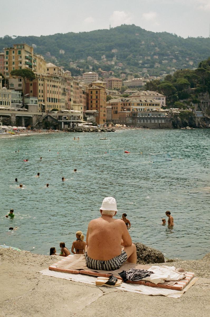 A man in a white hat sits on a concrete ledge overlooking a bay with swimmers and a town built into a hillside.