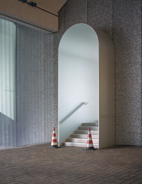 Two red and white traffic cones sit on a brick floor in front of an arched entryway with stairs and a handrail.
