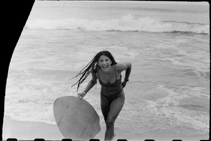 A woman with long hair and a swimsuit laughs while holding a skimboard at the beach.