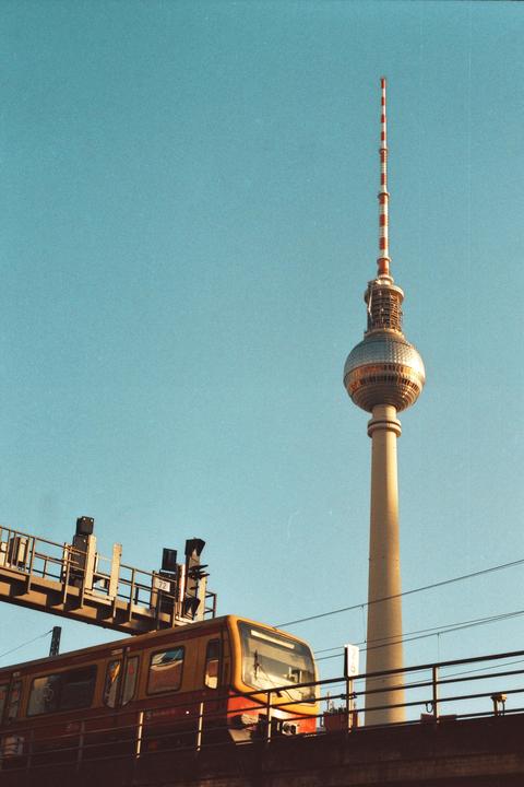 A yellow and red train passes in front of the Berlin TV Tower against a clear blue sky.