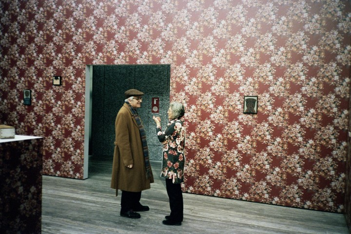 A man and a woman stand in a room with floral wallpaper, conversing near a doorway.