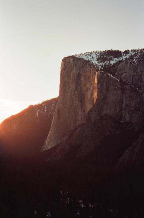 A large rock formation is illuminated by a golden light, with snow-capped trees visible on its peak and a dark forest below.