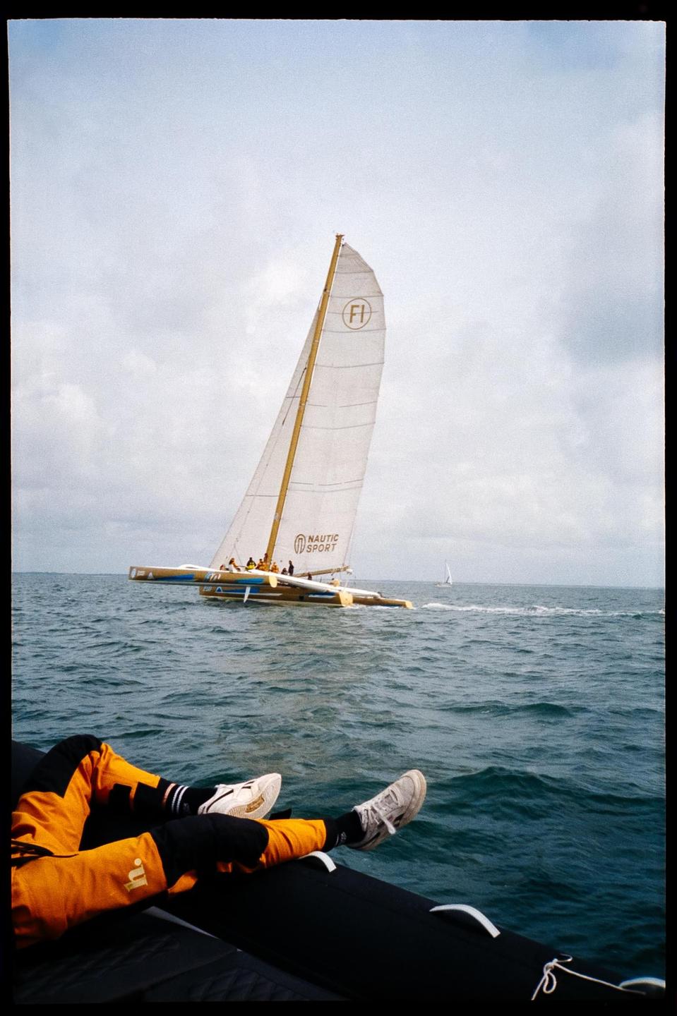 A person's legs are visible in the foreground of a body of water with a sailboat in the distance.
