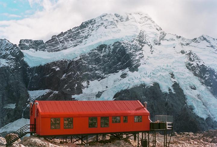 A red hut stands on stilts in front of a snow-covered mountain with a glacier.