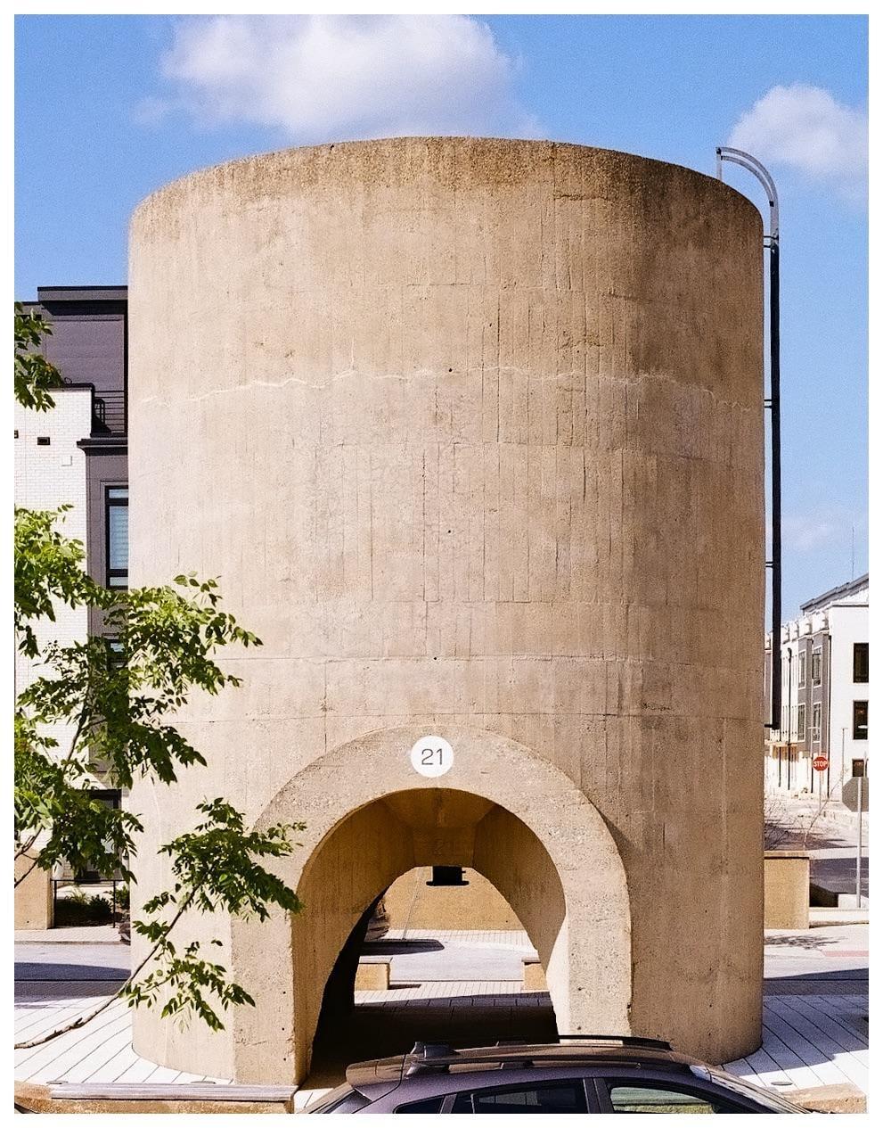 A large concrete cylinder with an arched opening stands next to a street with buildings and trees.