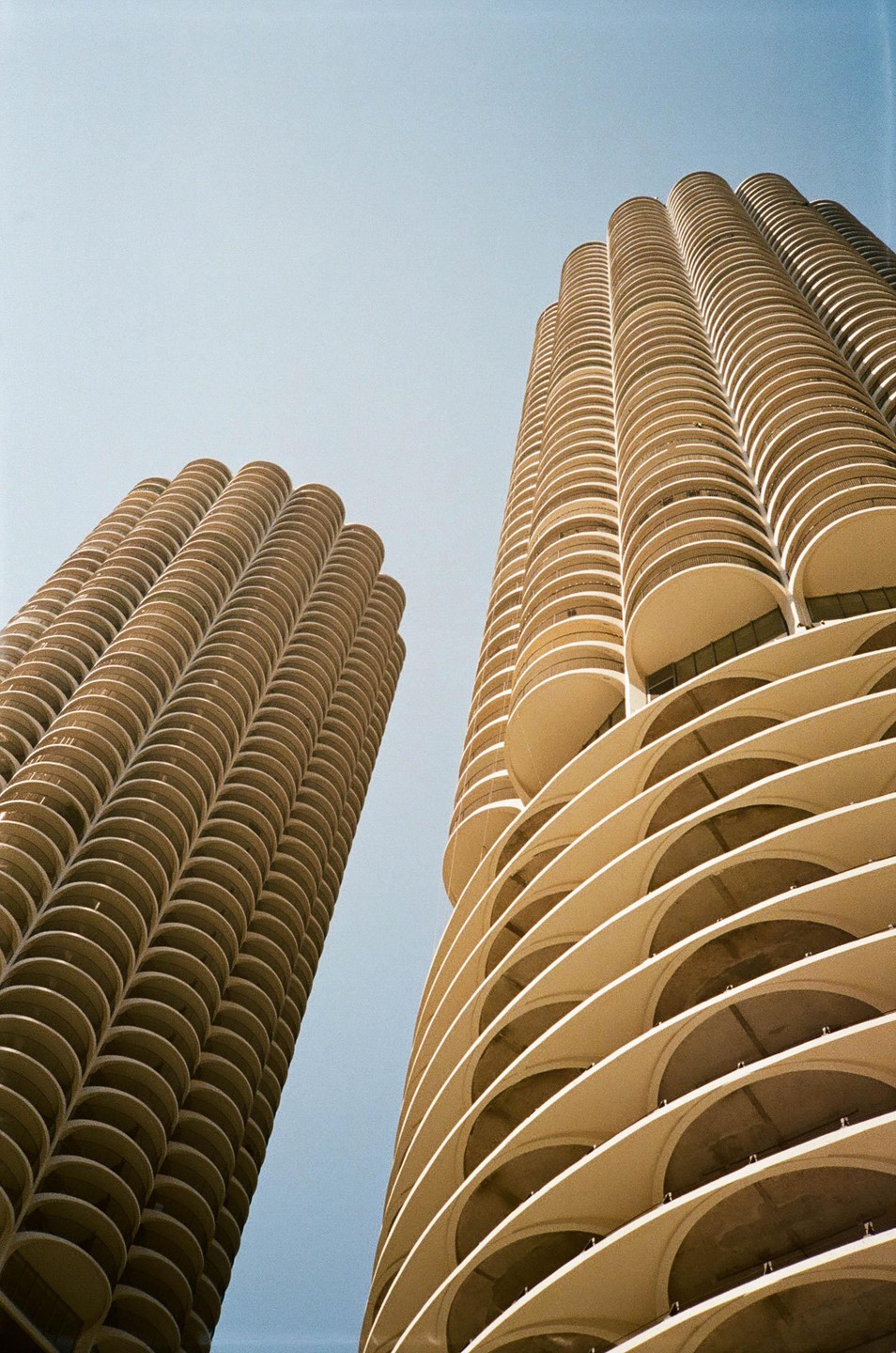 Two tall, cylindrical buildings with many balconies are photographed from below against a clear sky.