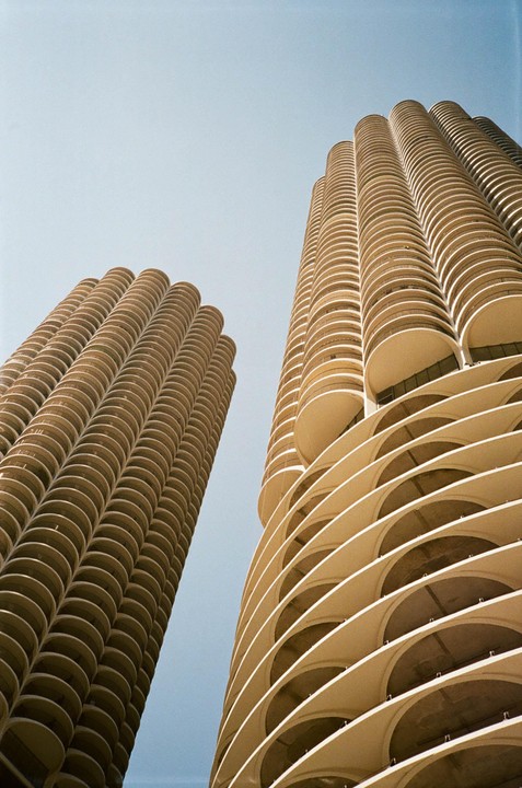 Two tall, cylindrical buildings with many balconies are photographed from below against a clear sky.