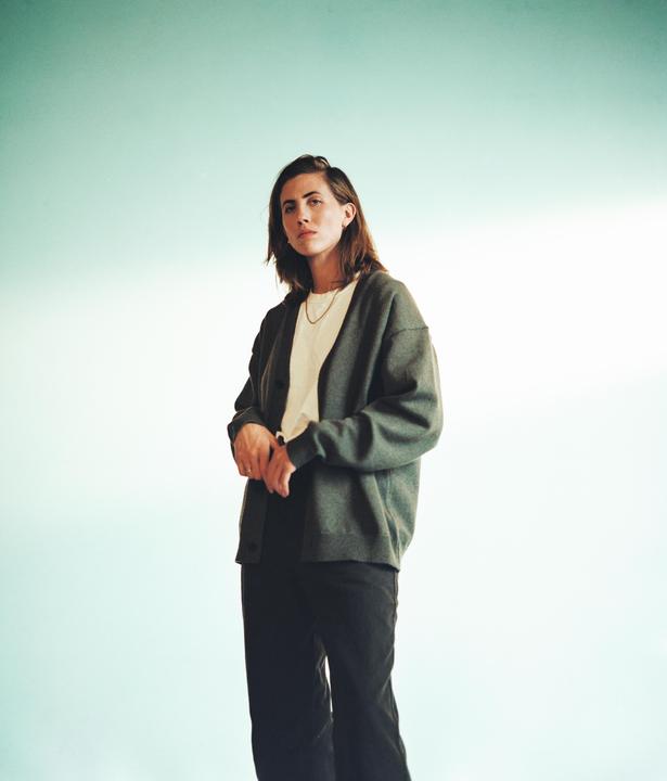A woman with dark hair stands against a light blue background, looking directly at the viewer.