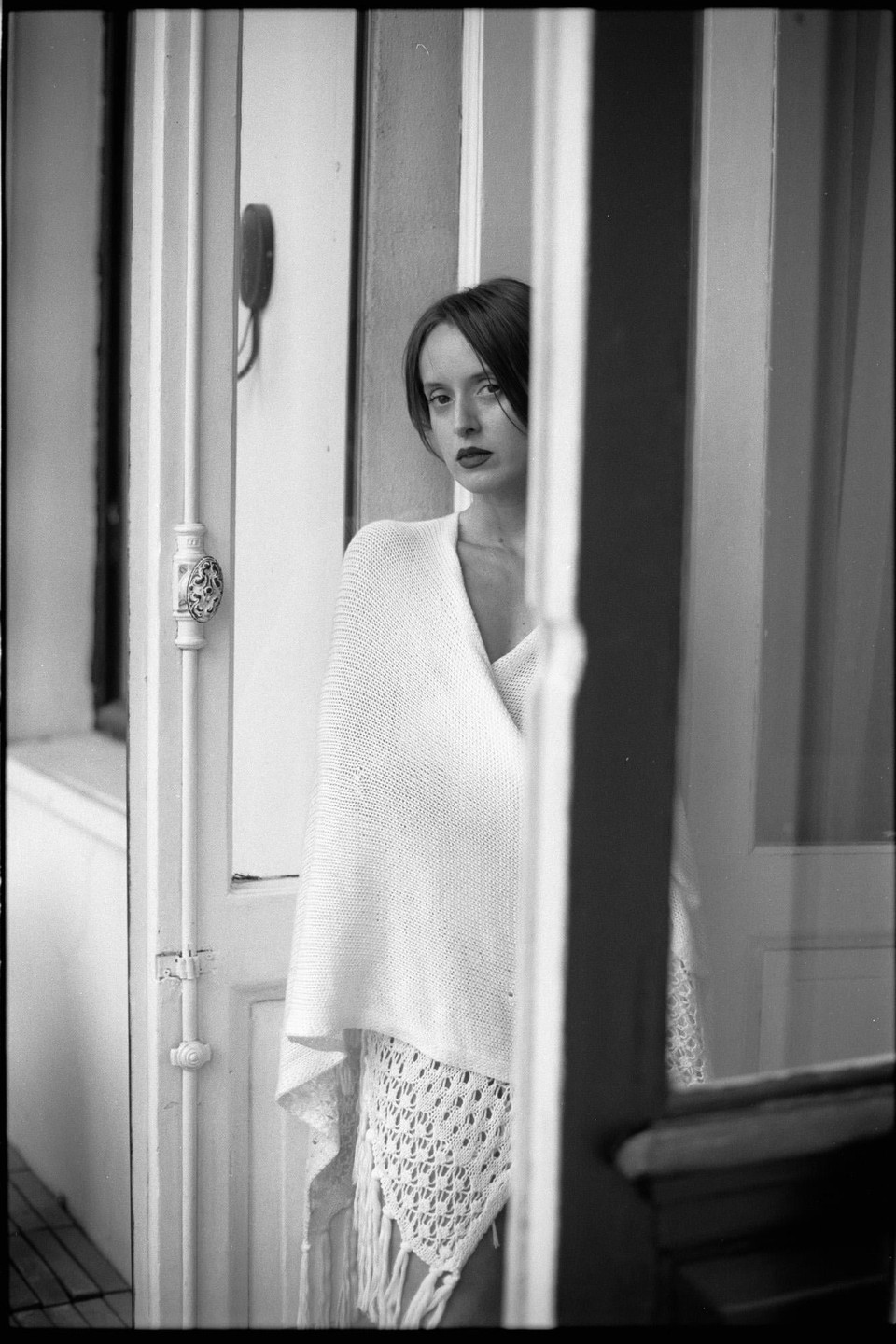 A woman with dark hair and red lipstick looks directly at the viewer while partially obscured by a doorway.