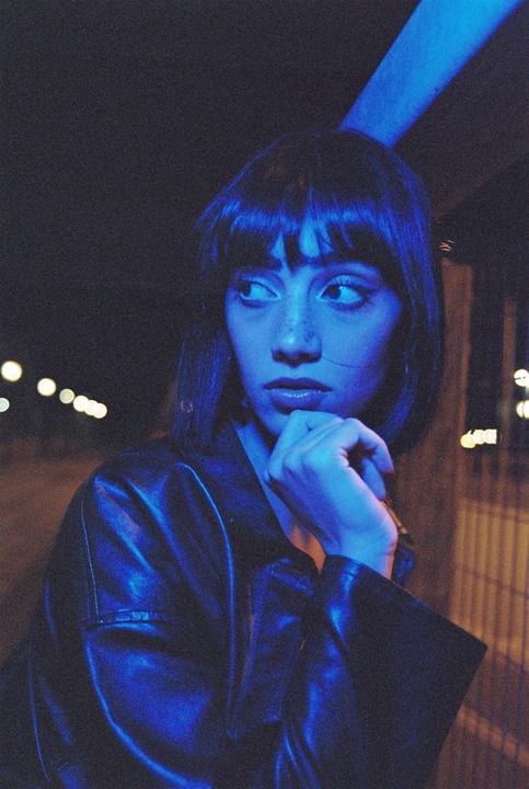 A woman with short hair and bangs looks to the side, illuminated by blue light at night.