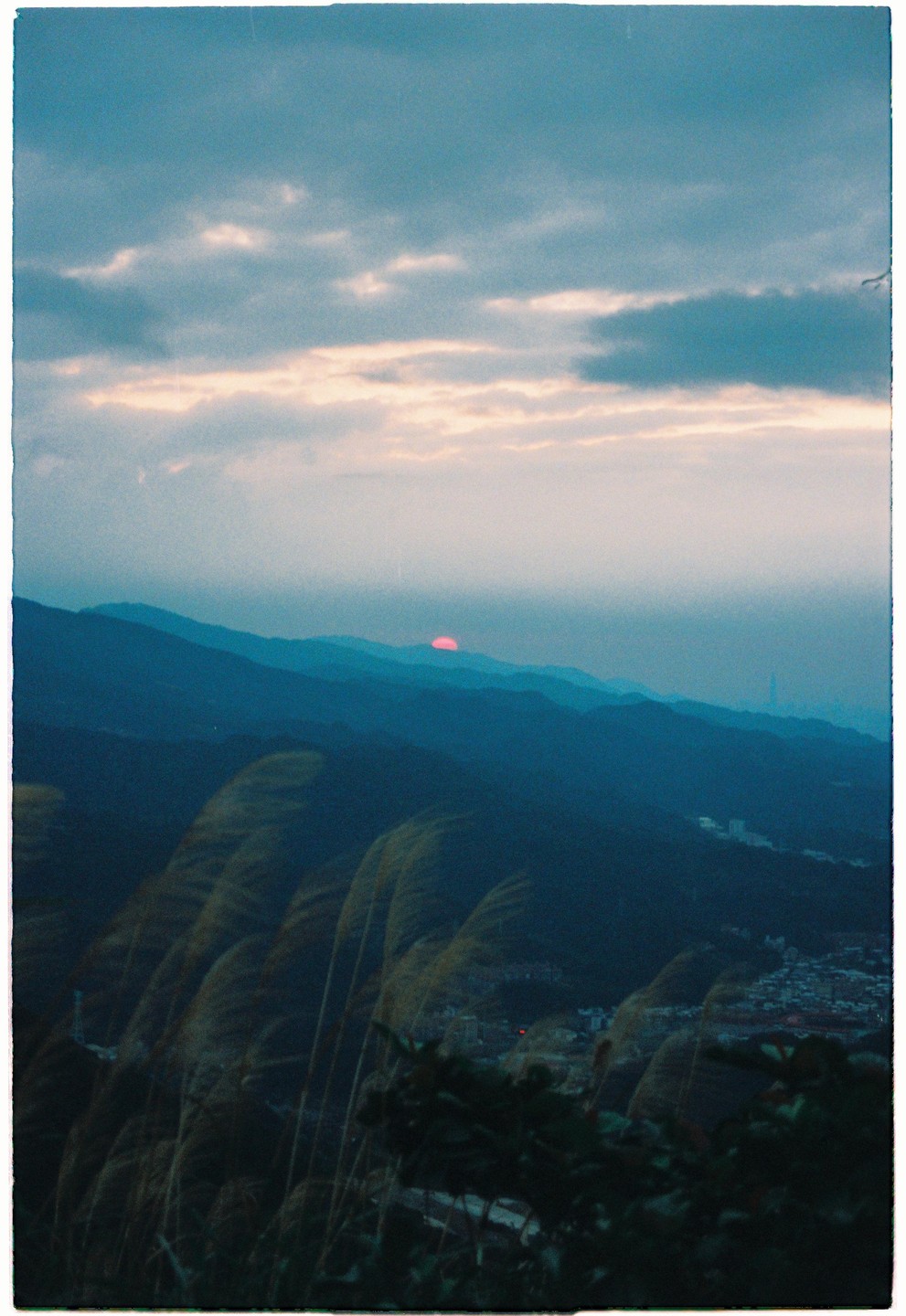 A red sun sets behind a range of mountains with tall grass in the foreground and a town in the valley.