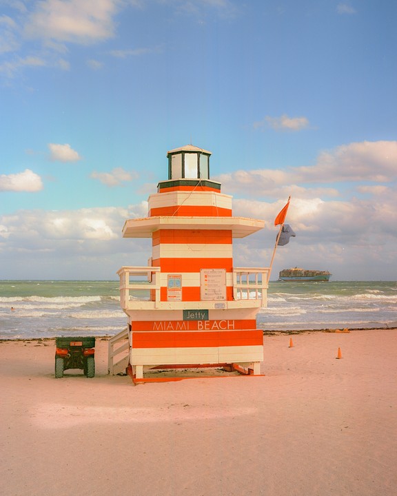 An orange and white striped lifeguard stand sits on a sandy beach with the ocean and a cargo ship in the background.