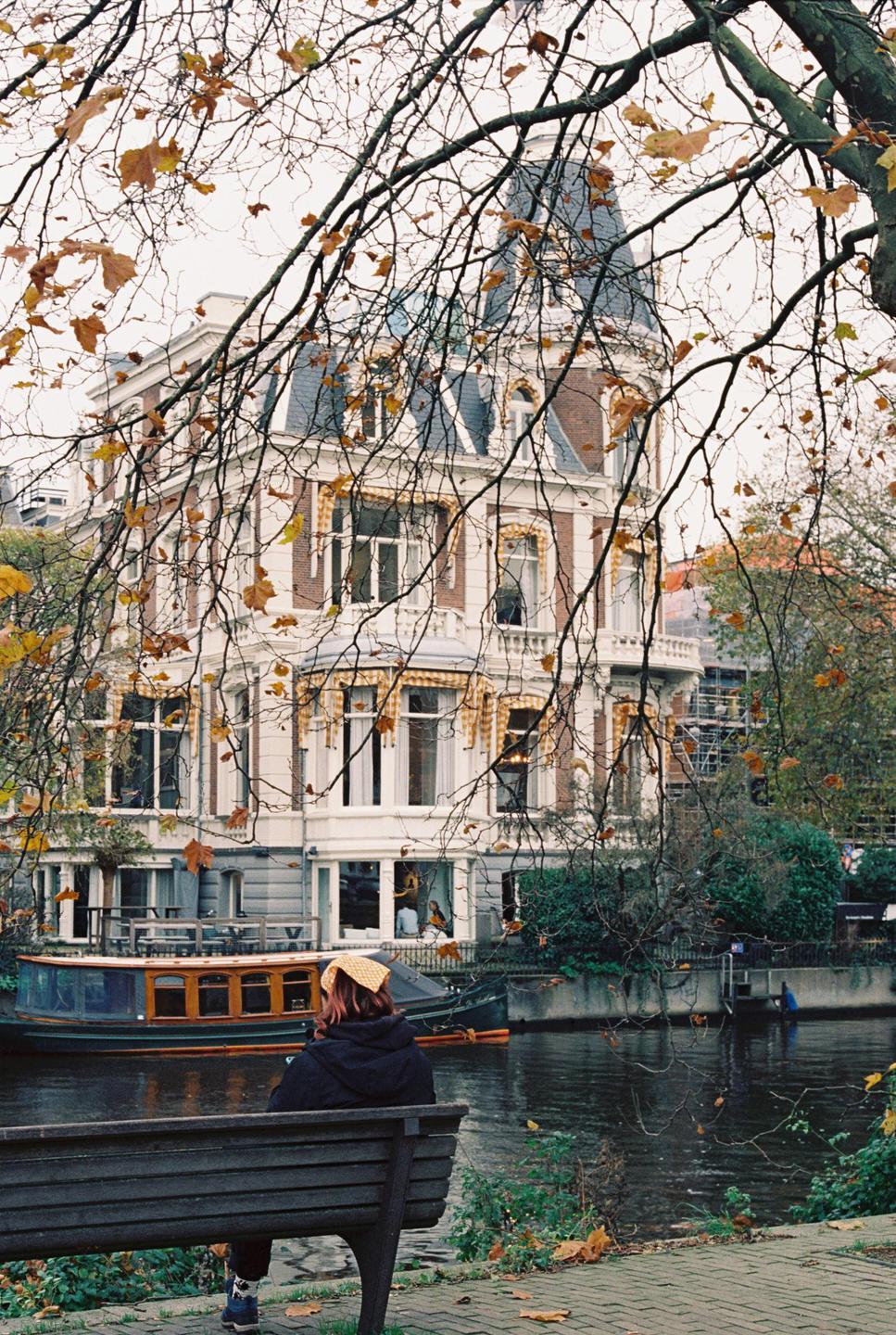 A person sits on a bench by a canal with a large building and a boat in the background.