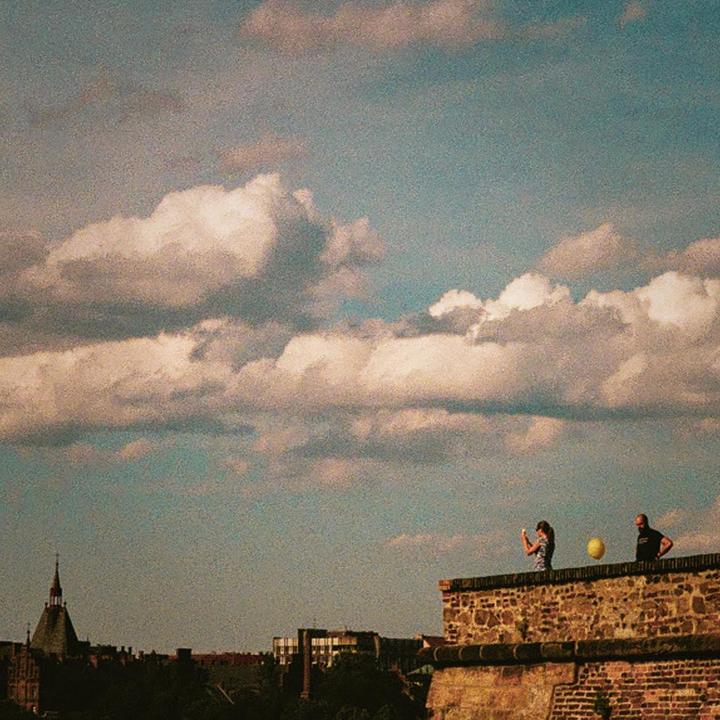 A woman and a man stand on a stone wall with a yellow balloon floating between them, overlooking a city.