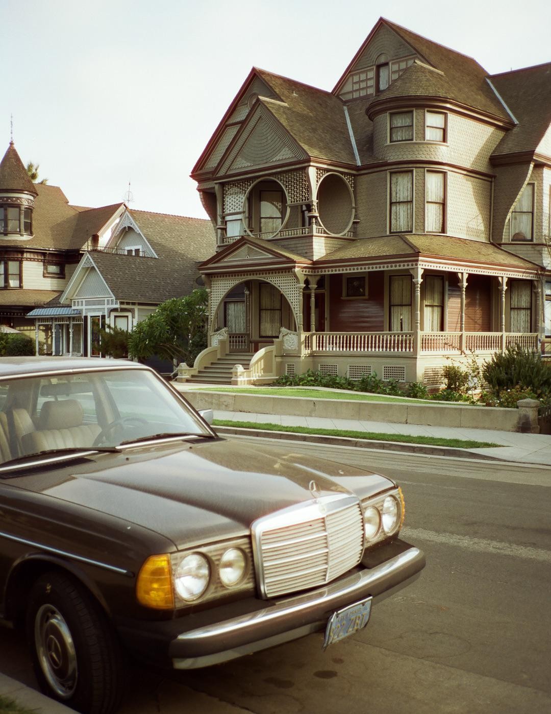 A brown car is parked on a street in front of a large, ornate house with a porch and steps.