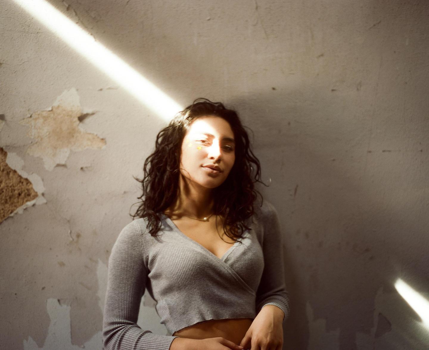 A woman with dark, curly hair and a gray top stands against a distressed wall with a beam of light across her face.