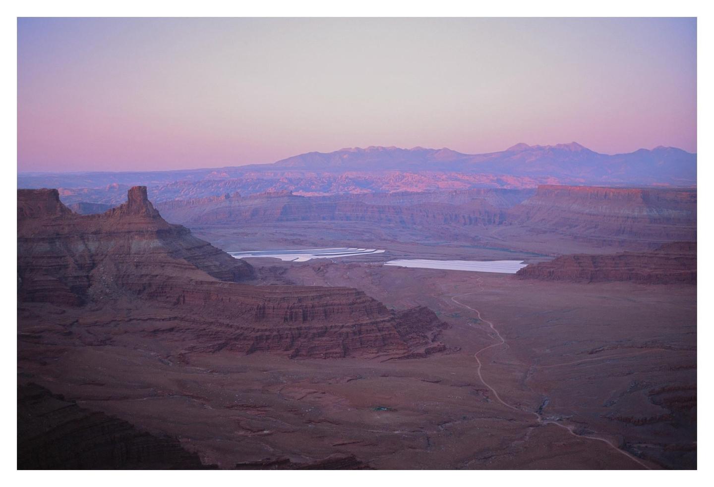 A vast canyon landscape with mesas and distant mountains is illuminated by the pink and purple light of dusk.