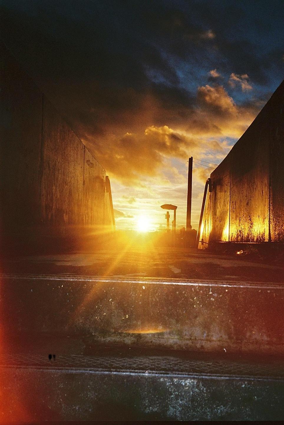 The sun sets between two tall structures, casting a bright flare across the frame.