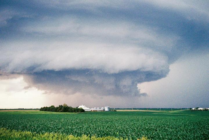 A large storm cloud looms over a green field with a farm in the distance.