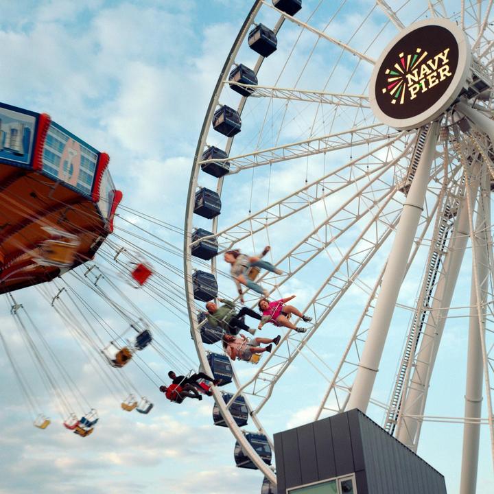 People ride a swing ride and a ferris wheel at an amusement park under a cloudy sky.