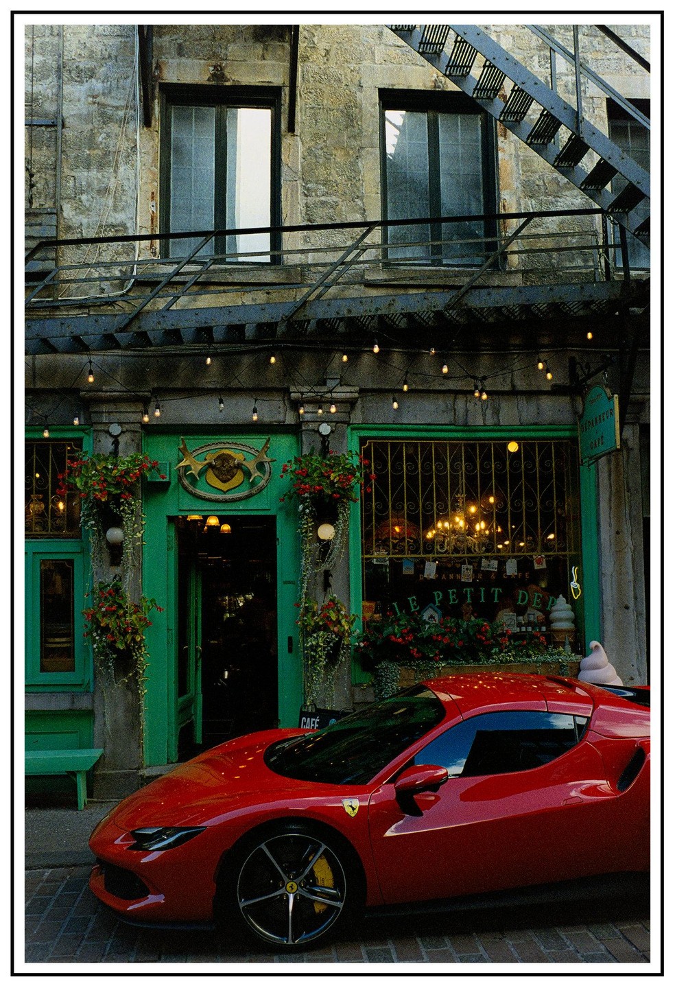 A red sports car is parked in front of a green cafe with string lights and window boxes.