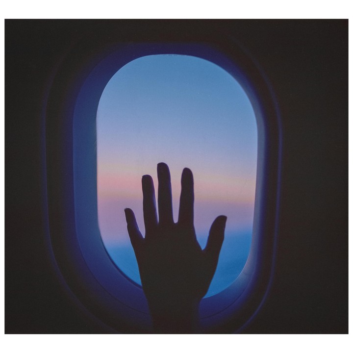 A hand is silhouetted against an airplane window with a colorful sky outside.