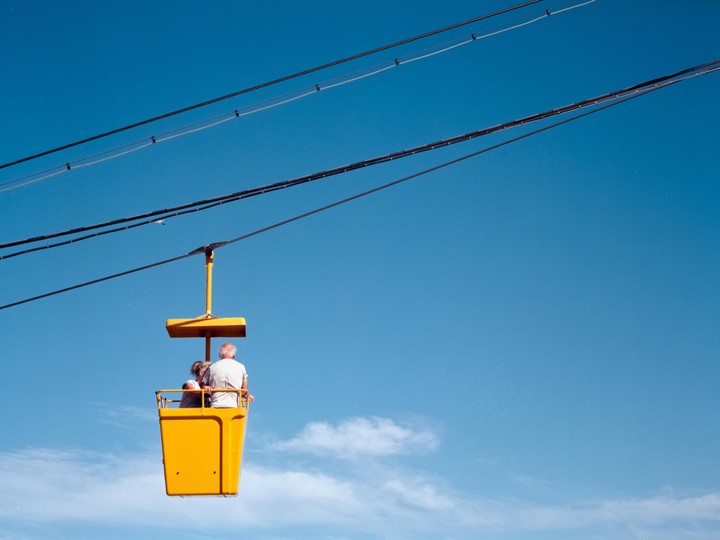 A man and a woman ride in a yellow gondola against a clear blue sky with power lines overhead.