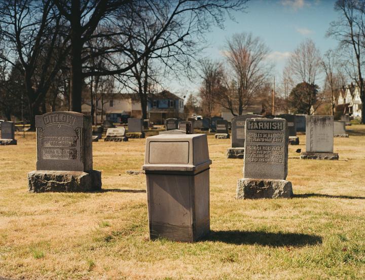 A trash can stands in the middle of a cemetery with gravestones and trees under a clear sky.