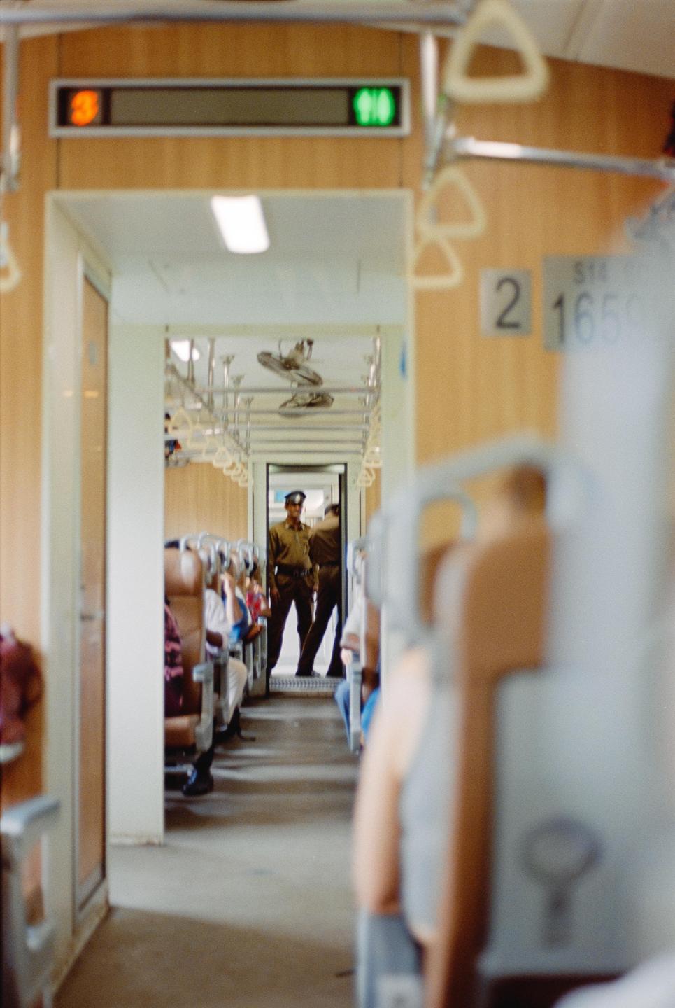 Two men in uniform stand in the aisle of a train car with passengers seated on both sides.