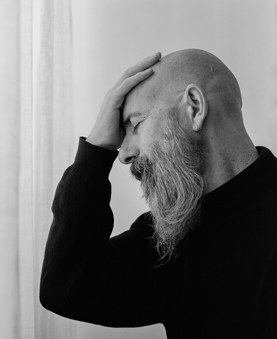 A man with a long beard and a shaved head holds his hand to his forehead.
