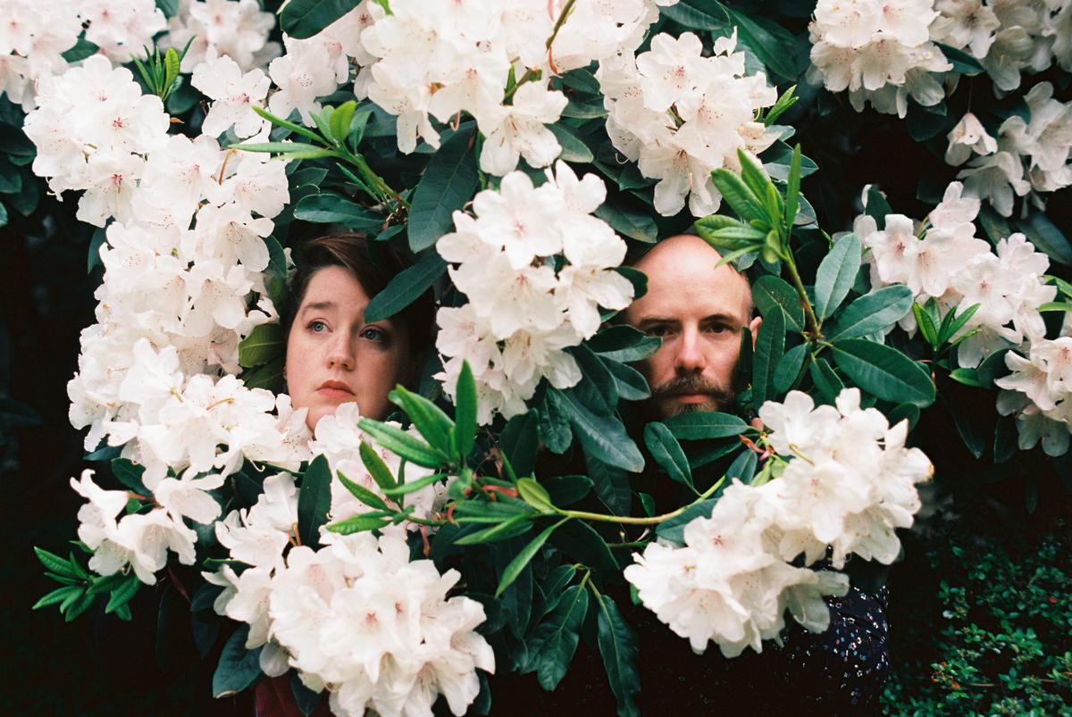 A woman and a man are partially hidden by white flowers and green leaves.