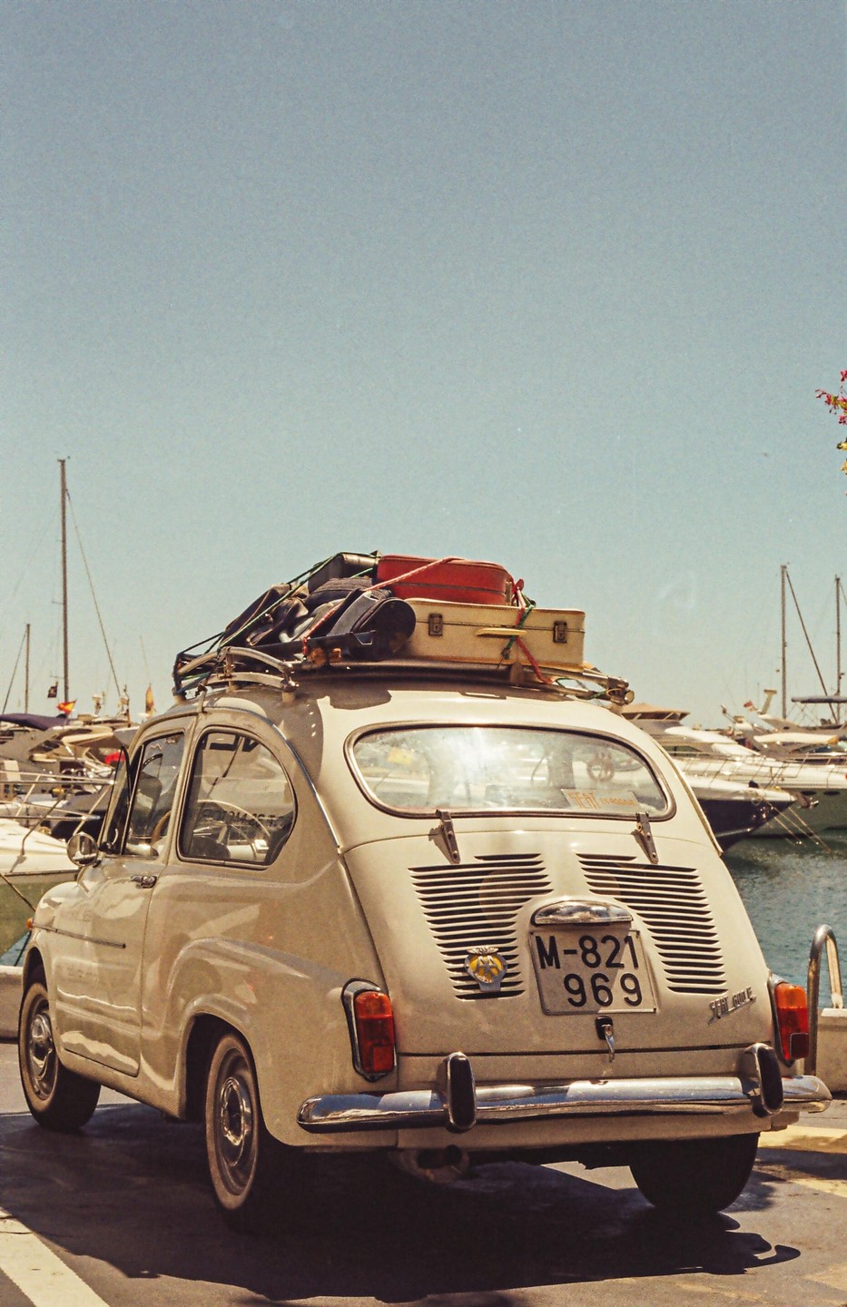 A small vintage car with luggage on its roof rack is parked next to a harbor filled with boats.