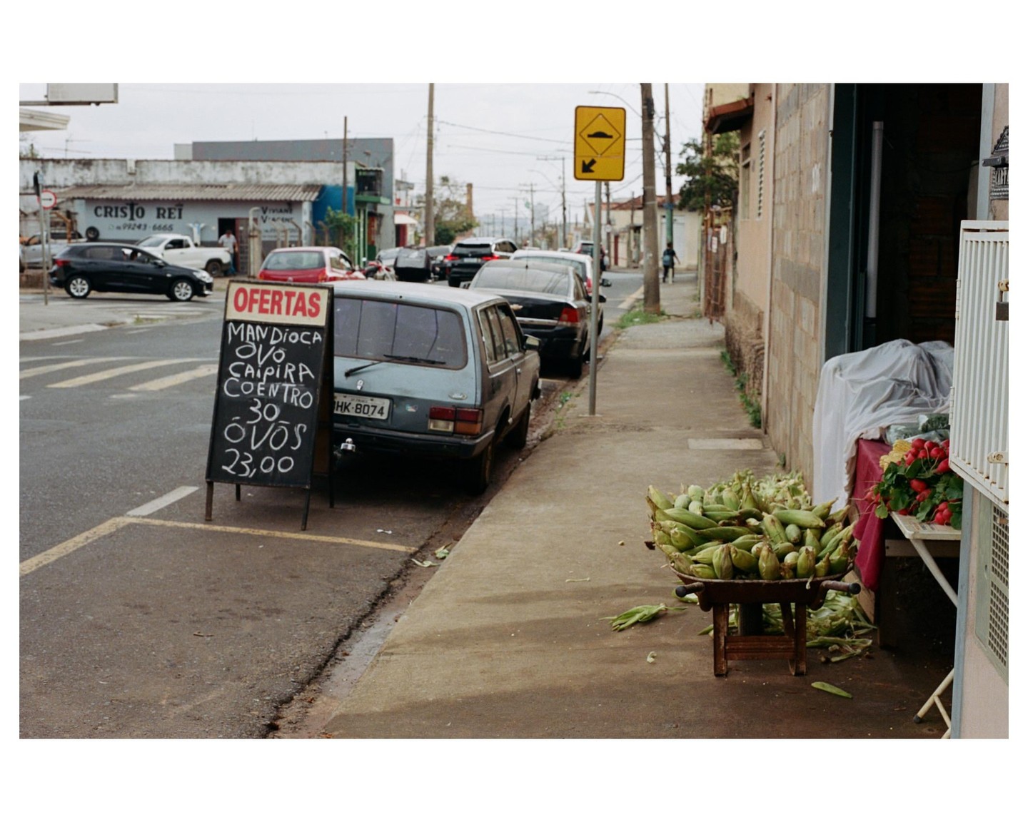 A street vendor's stall with corn and radishes sits on a sidewalk next to a parked car and a chalkboard sign advertising eggs.
