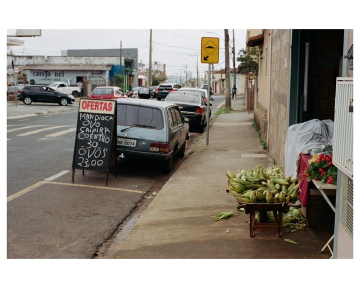 A street vendor's stall with corn and radishes sits on a sidewalk next to a parked car and a chalkboard sign advertising eggs.