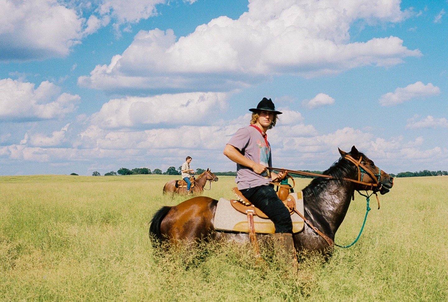 Two men ride horses through a tall grass field under a blue sky with clouds.