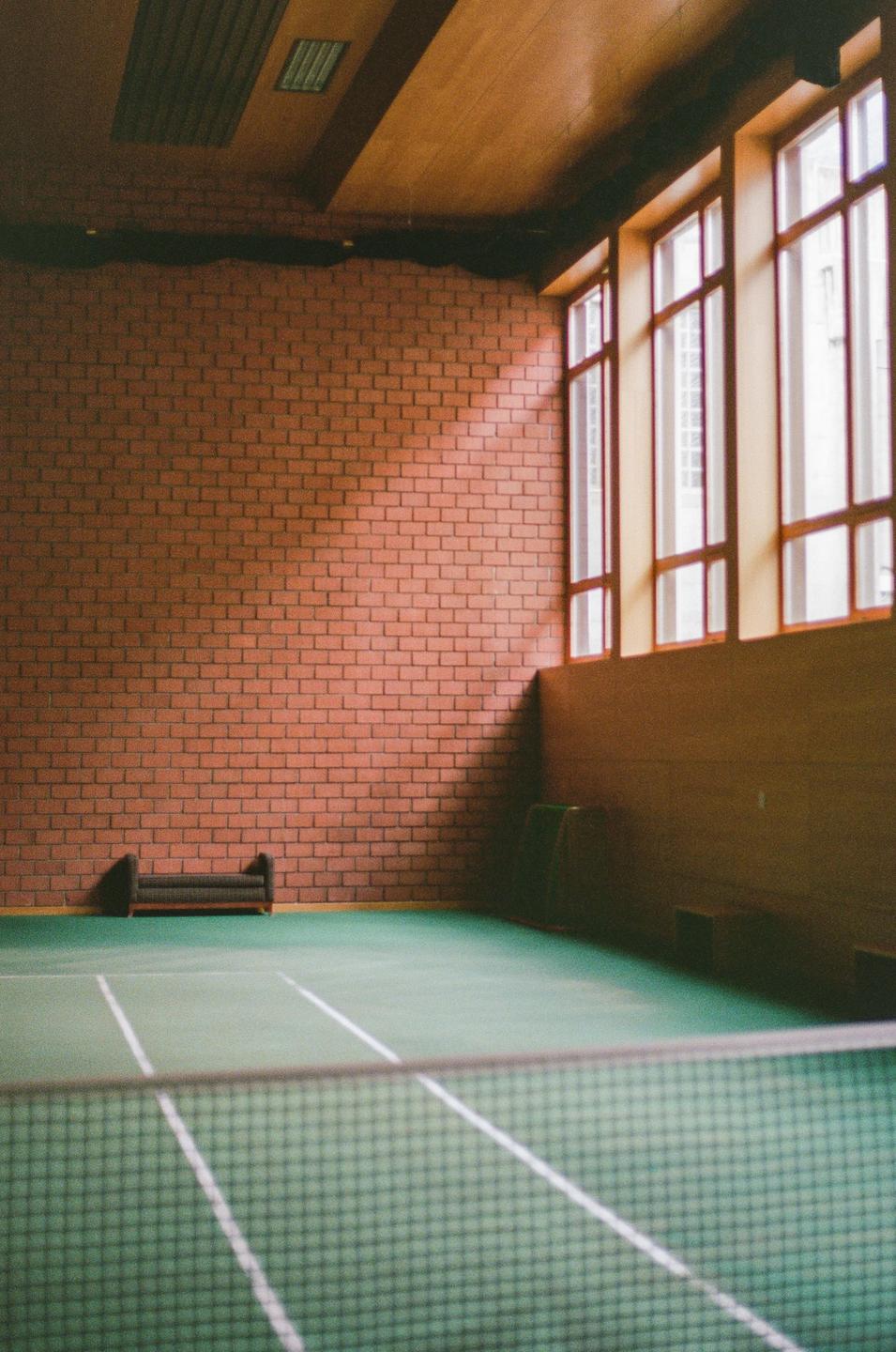 Sunlight streams through tall windows onto a brick wall and green court with a net in the foreground.
